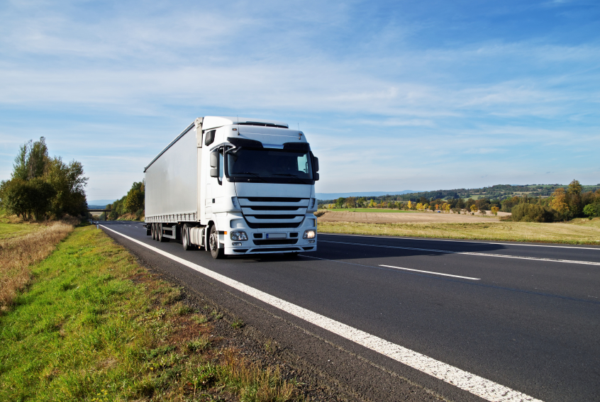 White truck travels on the asphalt road in the countryside. Fields, meadows and trees in early autumn colors in the background.